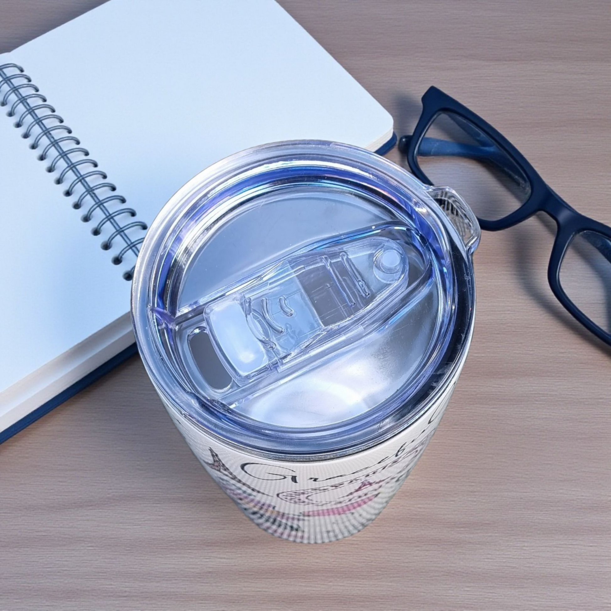 Clear tumbler with a lid on a wooden surface next to a notebook and glasses