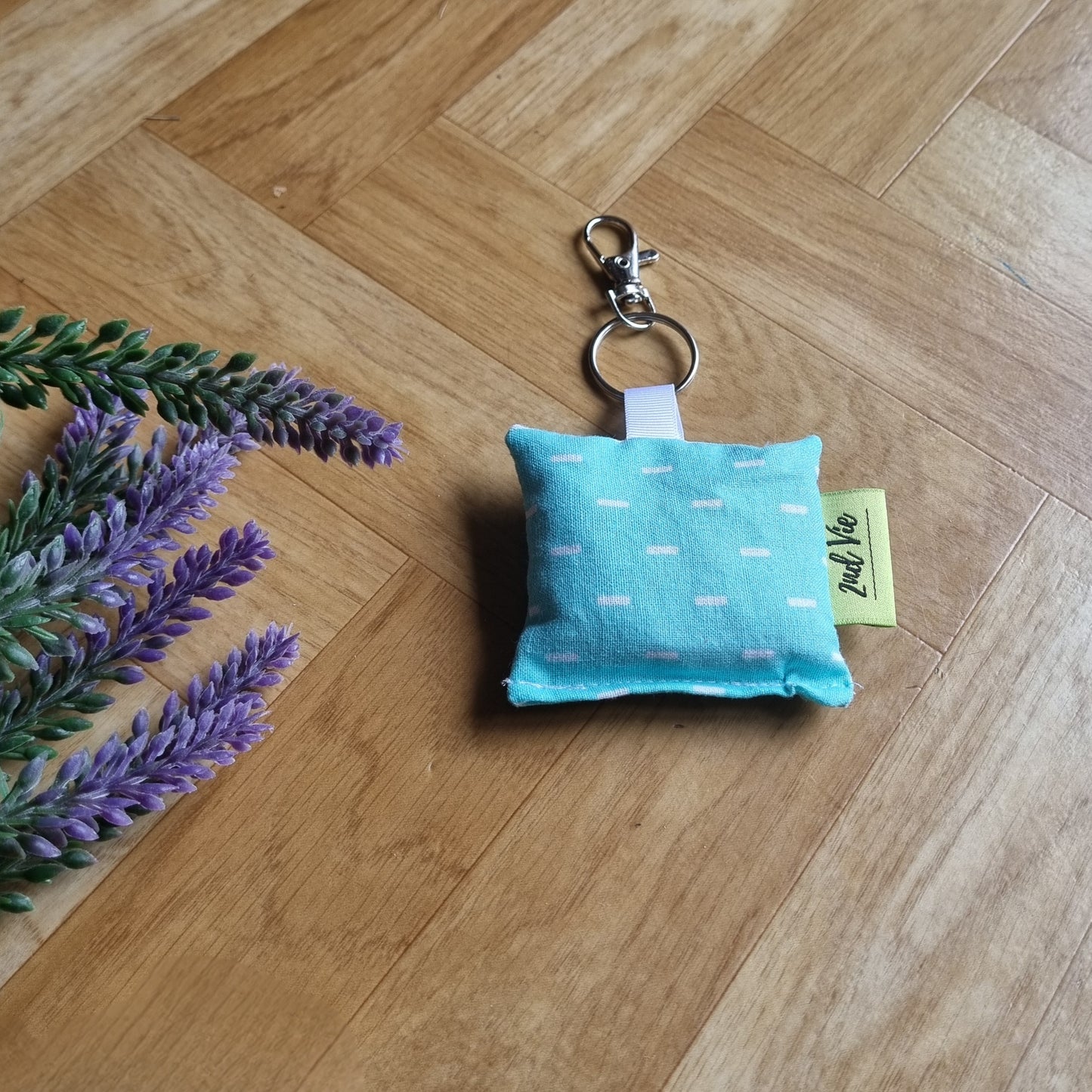 Turquoise keychain pillow with a tag on a wooden floor with lavender flowers.