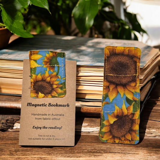 Sunflower-themed magnetic bookmark on a wooden surface with a plant in the background