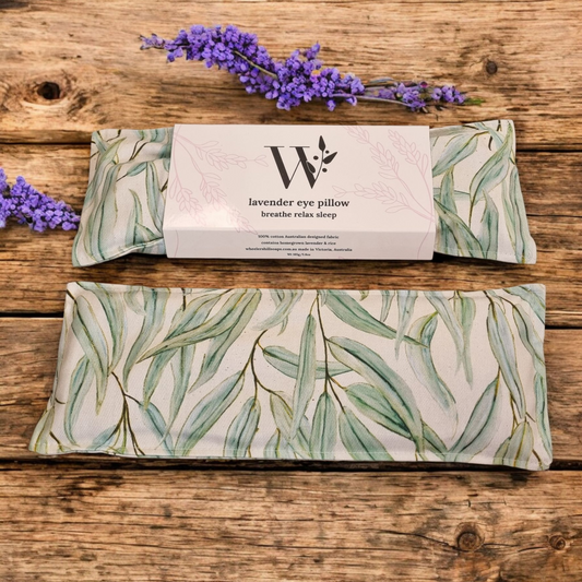 rectangular lavender eye pillows, featuring a light background with a subtle Australian flora design of eucalyptus leaves, are displayed on a warm, rustic wooden surface. A sprig of fresh lavender flowers is placed decoratively to the left and right of the pillows, enhancing the natural and calming aesthetic.
