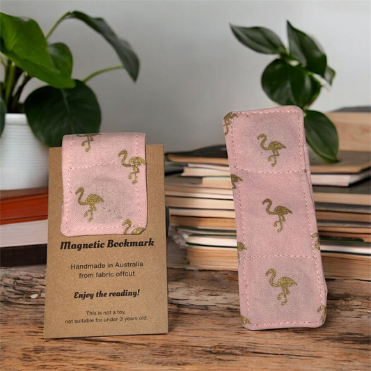 Two magnetic bookmarks with flamingo design on a wooden surface with books and plants in the background.